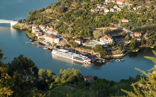 Flusskreuzfahrtschiff am Anleger in ländlicher Umgebung mit Häusern und Hügeln.