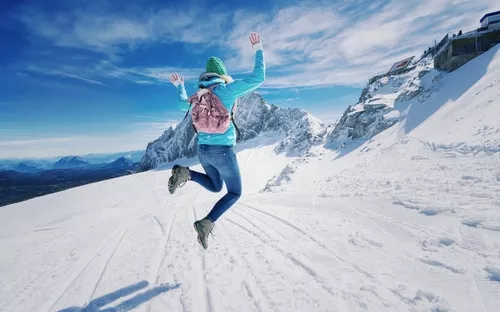 Person springt fröhlich im Schnee vor Bergkulisse.