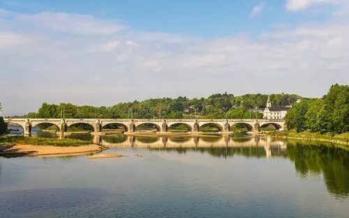 Fluss mit Brücke und bewaldeter Landschaft im Hintergrund unter blauem Himmel.