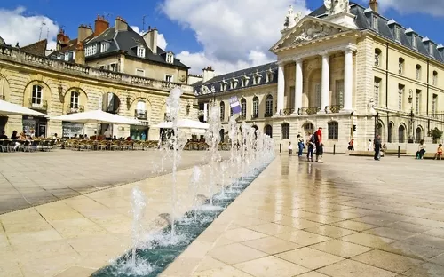 Place mit Springbrunnen und historischen Gebäuden bei Sonnenschein.
