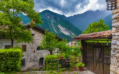 Steinhaus mit Garten und Tor vor bewaldetem Gebirgspanorama.