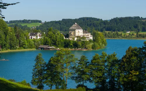 Schloss am malerischen See umgeben von Wald und Hügeln bei sonnigem Himmel.