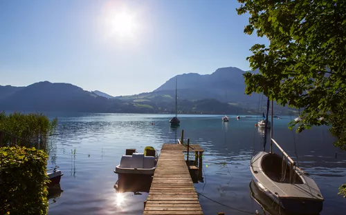 Steg am See mit Booten und Bergen im Hintergrund bei Sonnenschein.