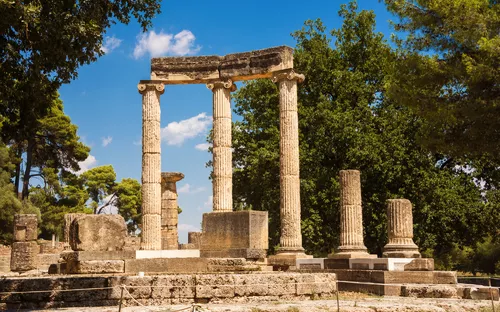 Ruinen antiker griechischer Säulen vor einem blauen Himmel und umgeben von Bäumen.