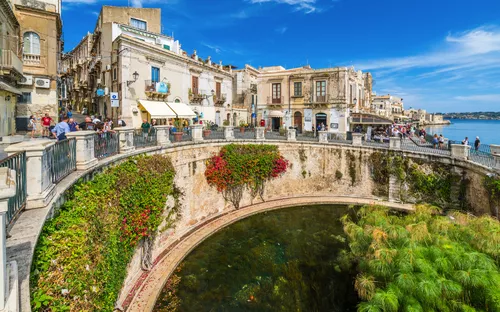Historische Gebäude mit Bogen und Pflanzen an einer Wasserquelle unter blauem Himmel.