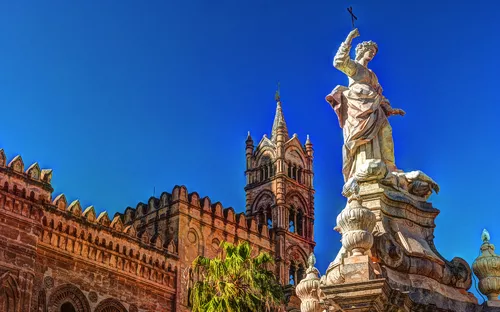 Statue und Kirche vor blauem Himmel, Palmen im Vordergrund.
