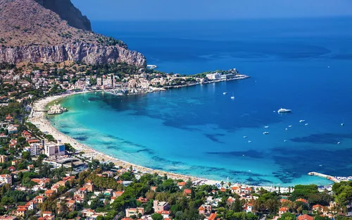 Küstenstadt mit Strand und blauem Meer an einer Bucht, umgeben von Bergen und Häusern.