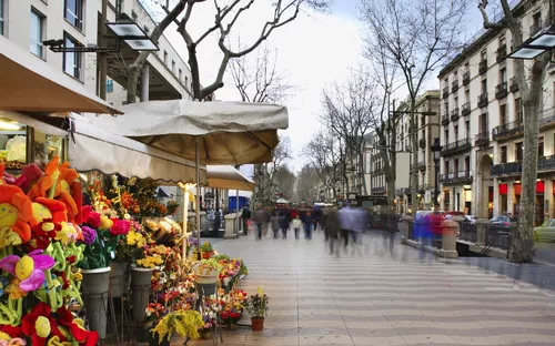 Blumenstand an einer belebten Straße mit Fußgängern und Bäumen im Hintergrund.