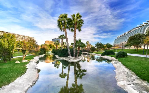 Palmeninsel in modernem Park mit Gebäuden im Hintergrund, reflektiert in einem Teich.