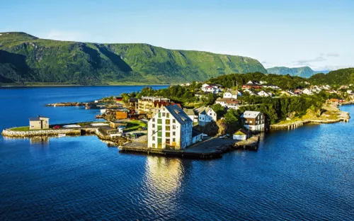 Luftaufnahme einer kleinen Küstenstadt an einem Fjord mit grünen Bergen im Hintergrund.
