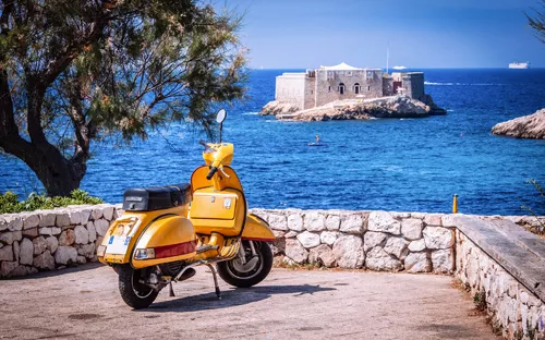 Gelber Motorroller vor Meer und Insel mit Festung im Hintergrund.