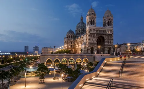 Basilika bei Dämmerung mit beleuchtetem Vorplatz und Treppe in urbaner Umgebung.
