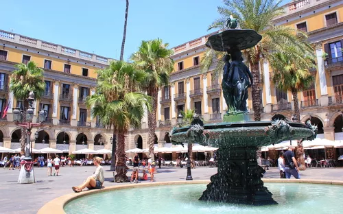 Brunnen auf einem Platz mit Palmen und Menschen im Hintergrund.