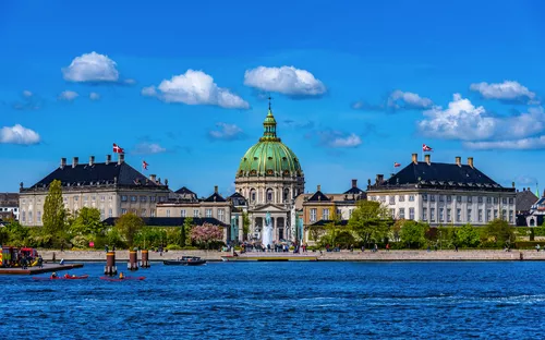 Kirche mit grünlicher Kuppel und Gebäuden am Wasser unter blauem Himmel.