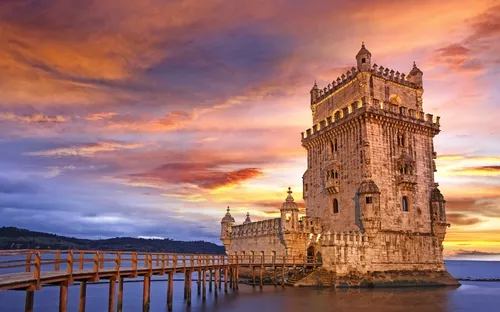 Ein historischer Turm an einem Flussufer bei Sonnenuntergang, umgeben von einem hölzernen Steg.