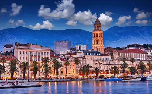 Historische Stadt am Meer mit Bergen und beleuchtetem Turm bei Dämmerung.