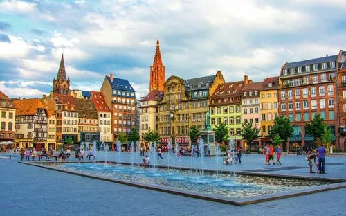 Platz mit Springbrunnen und historischen Gebäuden im Hintergrund bei bewölktem Himmel.