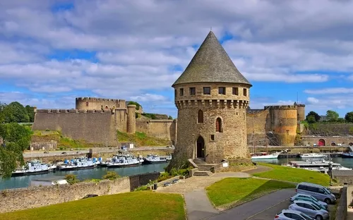 Mittelalterliche Burg mit Rundturm und Hafen im Vordergrund bei bewölktem Himmel.