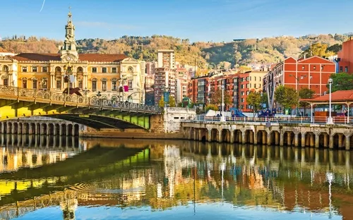 Historische Gebäude und Brücke am Flussufer, im Wasser reflektiert.