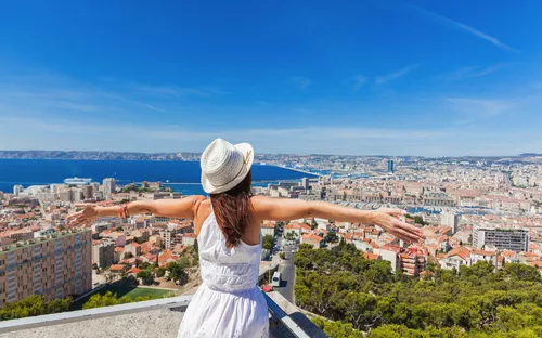 Frau in weißem Kleid steht mit ausgebreiteten Armen über einer Stadt mit Meerblick.
