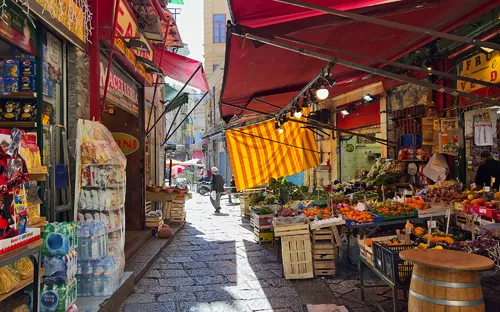 Marktstraße mit Obst- und Gemüseständen unter roten Sonnenschirmen.