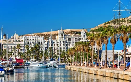 Hafenpromenade mit Yachten, Palmen und historischen Gebäuden unter blauem Himmel.