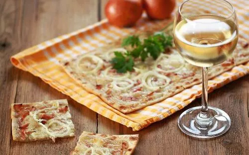 Flammkuchen und Weißwein auf einem Holztisch mit Zwiebeln und Petersilie.