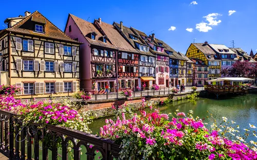Fachwerkhäuser und Blumen an einem Kanal unter blauem Himmel in einer europäischen Stadt.