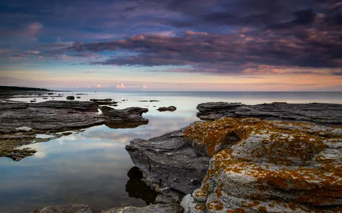Steinige Küste bei Sonnenuntergang mit Wolken und ruhigem Wasser.