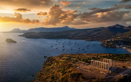 Küste bei Sonnenuntergang mit Tempel, Segelbooten und Bergen im Hintergrund.