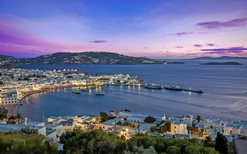 Hafenstadt bei Sonnenuntergang mit Meer, Gebäuden und Hügeln im Hintergrund.