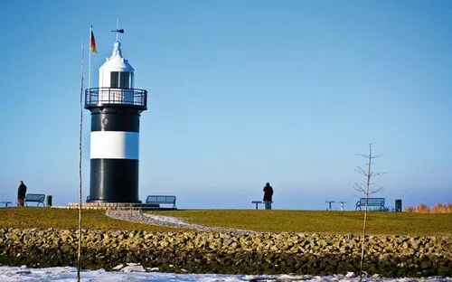 Schwarz-weißer Leuchtturm auf Wiese mit zwei Spaziergängern, blauem Himmel und Schnee.