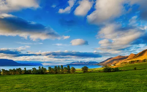 Grüne Wiese vor Bergen und See unter bewölktem Himmel