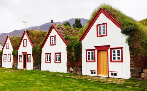 Drei weiße Holzhäuser mit Grasdächern in einer Berglandschaft.