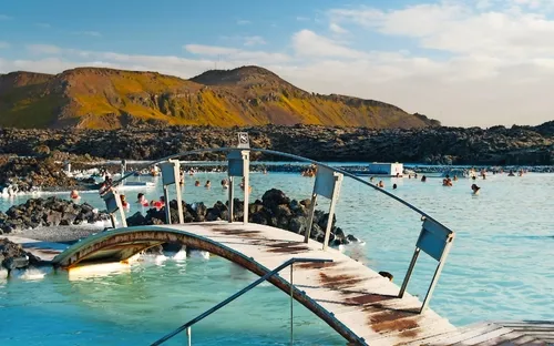 Natürliche heiße Quelle mit Badenden und Brücke, umgeben von Vulkanlandschaft.