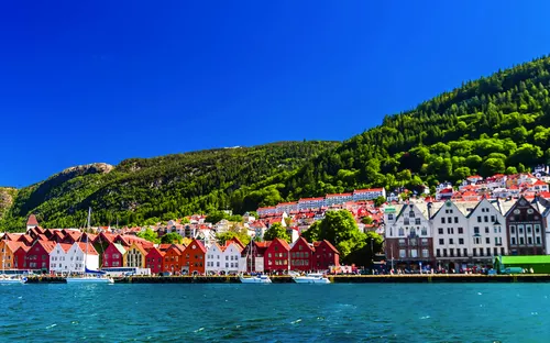 Häuserreihe am Wasser mit Bergen im Hintergrund bei klarem Himmel.