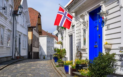 Gepflasterte Gasse mit weißen Holzhäusern und flaggendekorierten Eingängen.