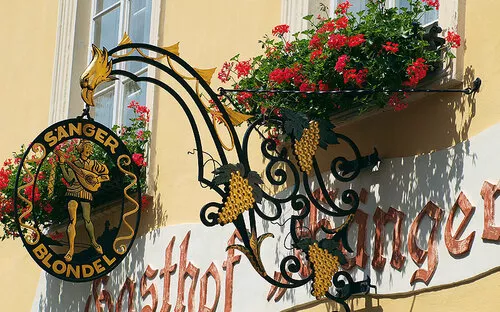 Eisernes Wirtshausschild mit Musikermotiv und Blumenkasten an gelber Hausfassade.