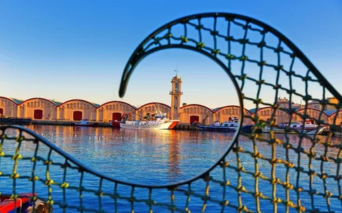 Hafen mit Uhrturm und Booten hinter einem Metallgitter bei Sonnenuntergang.