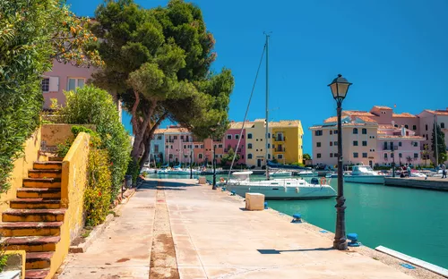 Promenade mit Booten im Yachthafen, umgeben von farbigen Gebäuden und Bäumen.