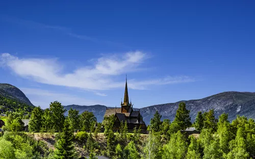 Historische Stabkirche in einer grünen Landschaft vor Bergkulisse und blauem Himmel.