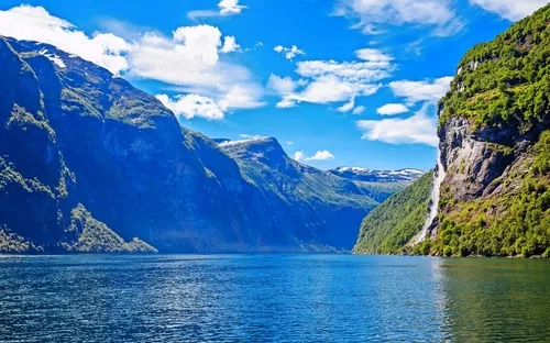 Blick auf einen Fjord mit Wasserfall, umgeben von bewaldeten Bergen unter blauem Himmel