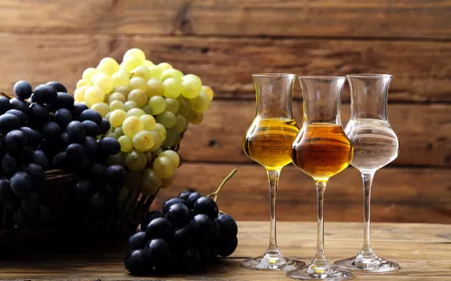 Drei Gläser Spirituosen und Trauben auf Holztisch vor Holzwand.