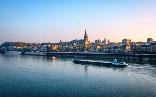 Stadtansicht mit Fluss, Kirchturm und Schiff im Vordergrund bei Sonnenaufgang.