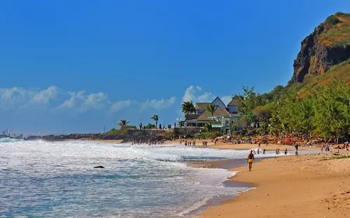 Strand mit Menschen, Meer und grüner Klippe unter klarem, blauem Himmel.