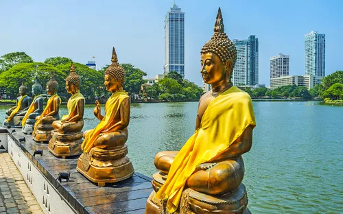 Reihe von Buddha-Statuen an einem See mit Hochhäusern im Hintergrund.
