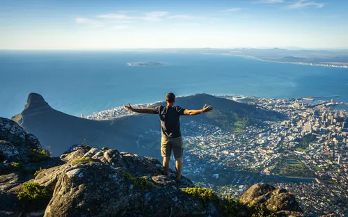Mann steht mit ausgebreiteten Armen auf Berg, blickt auf Küste und Stadt.