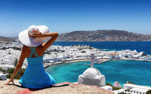 Frau in blauem Kleid und Hut blickt auf Küstenstadt mit blauem Meer und Hügel im Hintergrund.