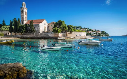 Küste mit Kloster, Türmen und Anlegestelle bei klarem, türkisfarbenem Meer.