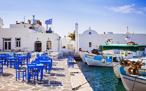 Hafenpromenade mit weißen Gebäuden, blauen Stühlen und Booten im Wasser